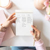 hands holding written on notepad with pink trim tape, desk, gold pen, blush planner, candle, pink flowers and vase