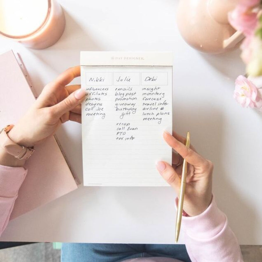 hands holding written on notepad with pink trim tape, desk, gold pen, blush planner, candle, pink flowers and vase