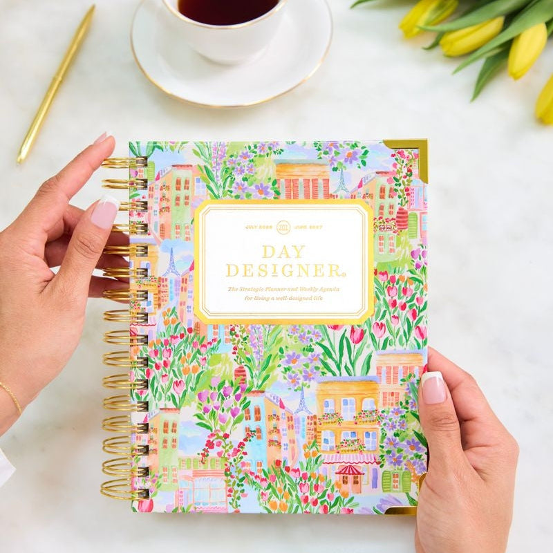 A person holds a colorful spiral-bound planner titled 'DAY DESIGNER' with a gold-accented cover. The scene is styled on a light marble surface with a cup of tea, yellow tulips, and a gold pen.