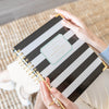 A person holding a spiral-bound Day Designer notebook with black and white horizontal stripes and gold rings, photographed from above on a woven rug.