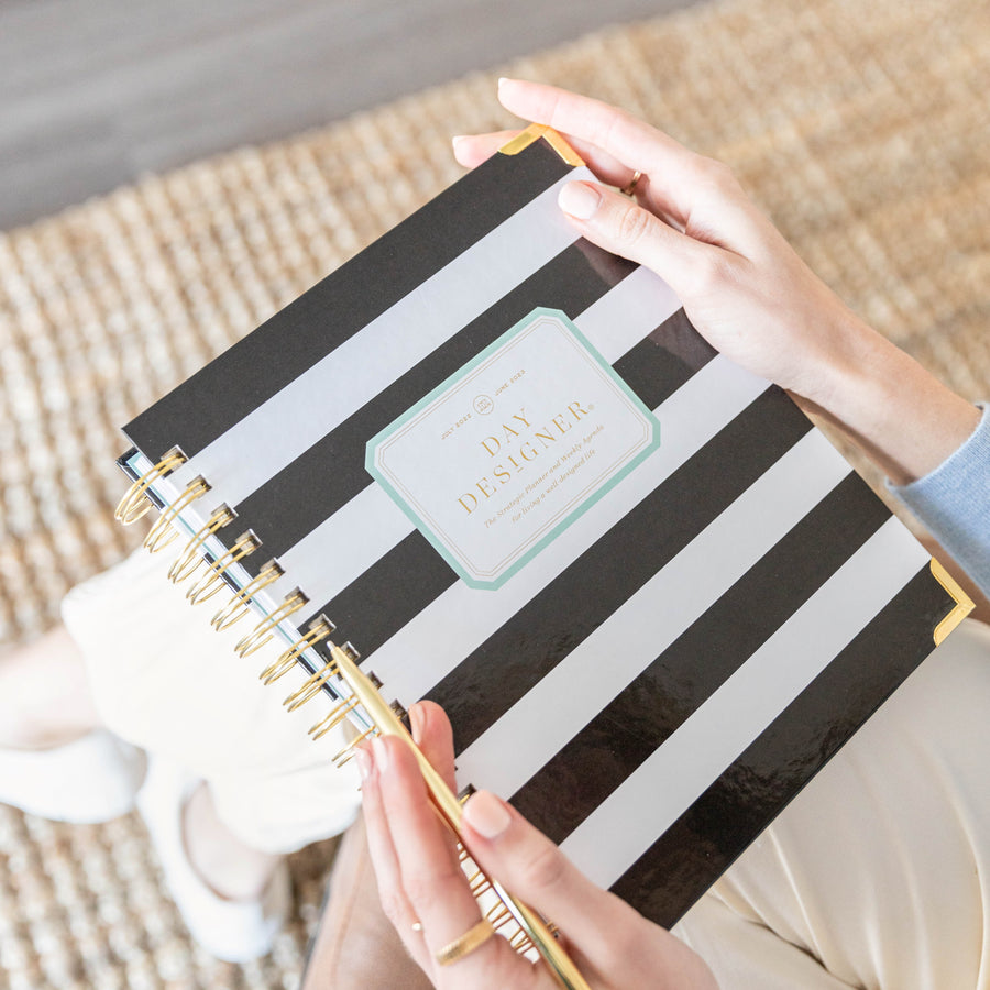 A person holding a spiral-bound Day Designer notebook with black and white horizontal stripes and gold rings, photographed from above on a woven rug.