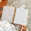 weekly open book planner, written on pages, gold binding and tabs, floral dress, hands, white pen, grey rug, wood table