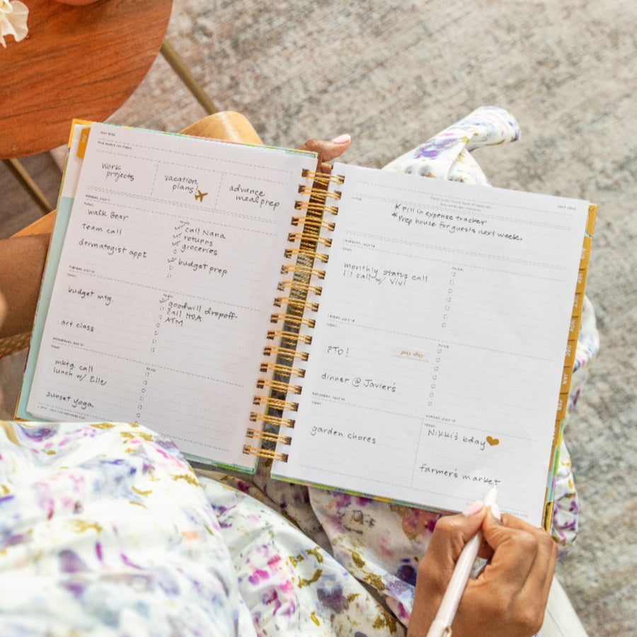 weekly open book planner, written on pages, gold binding and tabs, floral dress, hands, white pen, grey rug, wood table