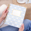 A person holds a spiral-bound planner with a white cover decorated with a light blue scallop pattern and a gold border. The scene is styled with blue jeans, a striped shirt, a woven round table, a cup of coffee, and a gold pen.