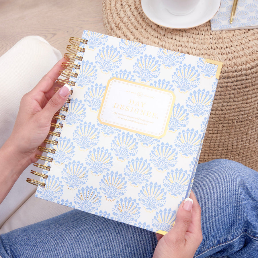 A person holds a spiral-bound planner with a white cover decorated with a light blue scallop pattern and a gold border. The scene is styled with blue jeans, a striped shirt, a woven round table, a cup of coffee, and a gold pen.