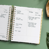 daily planner, open book, written on pages, gold binding and tabs, green background, palm leaf, brown dish