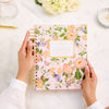 Floral planner held by hands on a light surface with flowers and a glass in the background