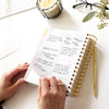 open book planner, written on page, gold binding and pen, candle, flowers, eye glasses, keyboard on white desk