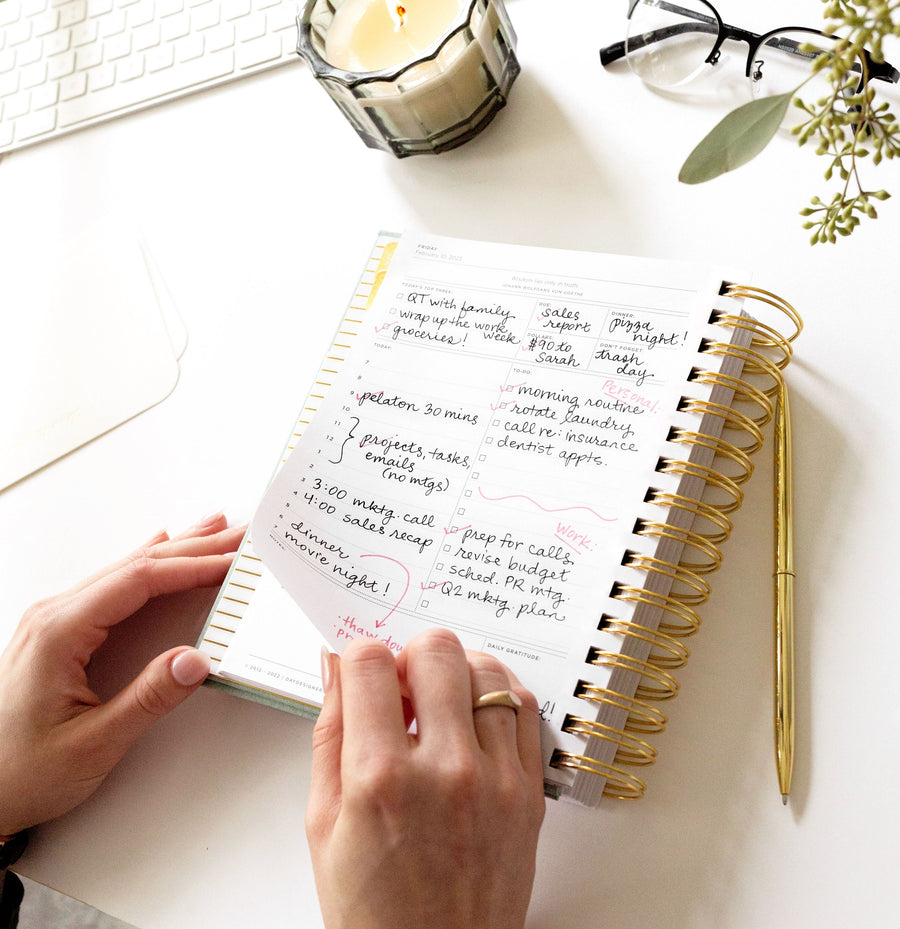 open book planner, written on page, gold binding and pen, candle, flowers, eye glasses, keyboard on white desk