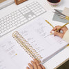 open planner book, written on pages, hands, gold binding, tabs, pen, and scissors, gray fabric, tea cup, white keyboard on white desk