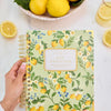 Hand holding a notebook with lemon design, surrounded by lemons on a white surface