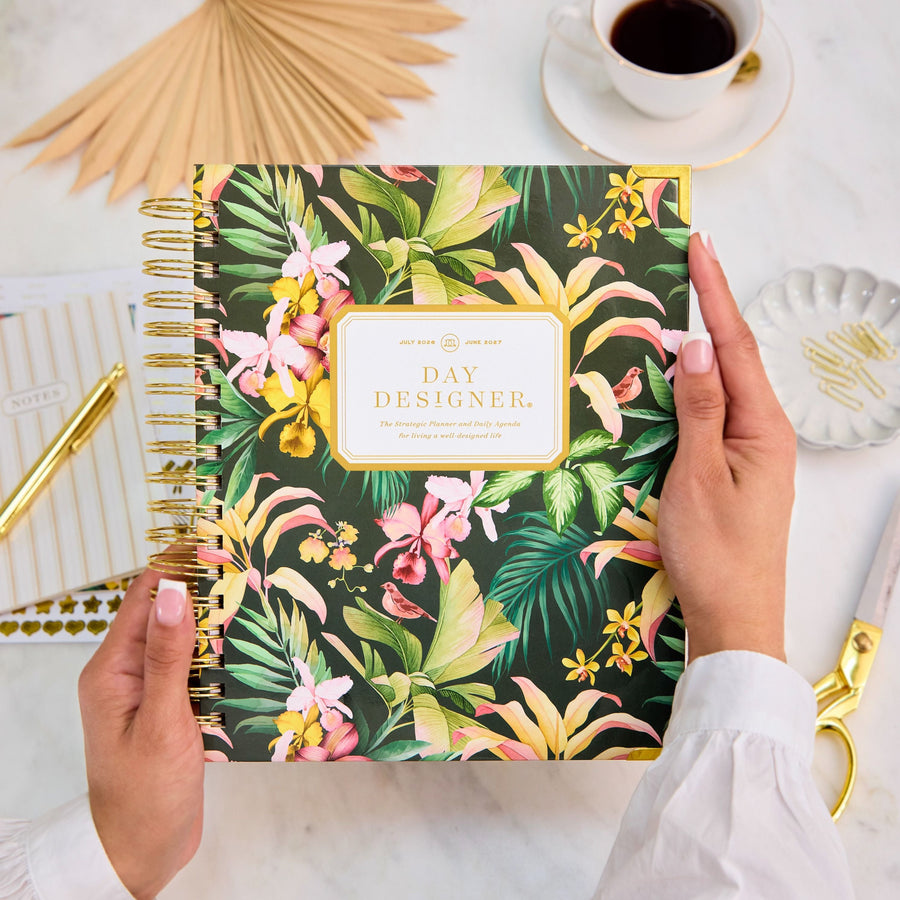 Floral notebook with 'Day Designer' branding held by a person on a desk with coffee and stationery.