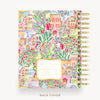 A spiral-bound notebook or planner shown from the back, featuring a colorful illustrated pattern of flowers, tulips, and small buildings. The cover has a cream label area with text, and the pages are bound with gold-tone spiral rings.