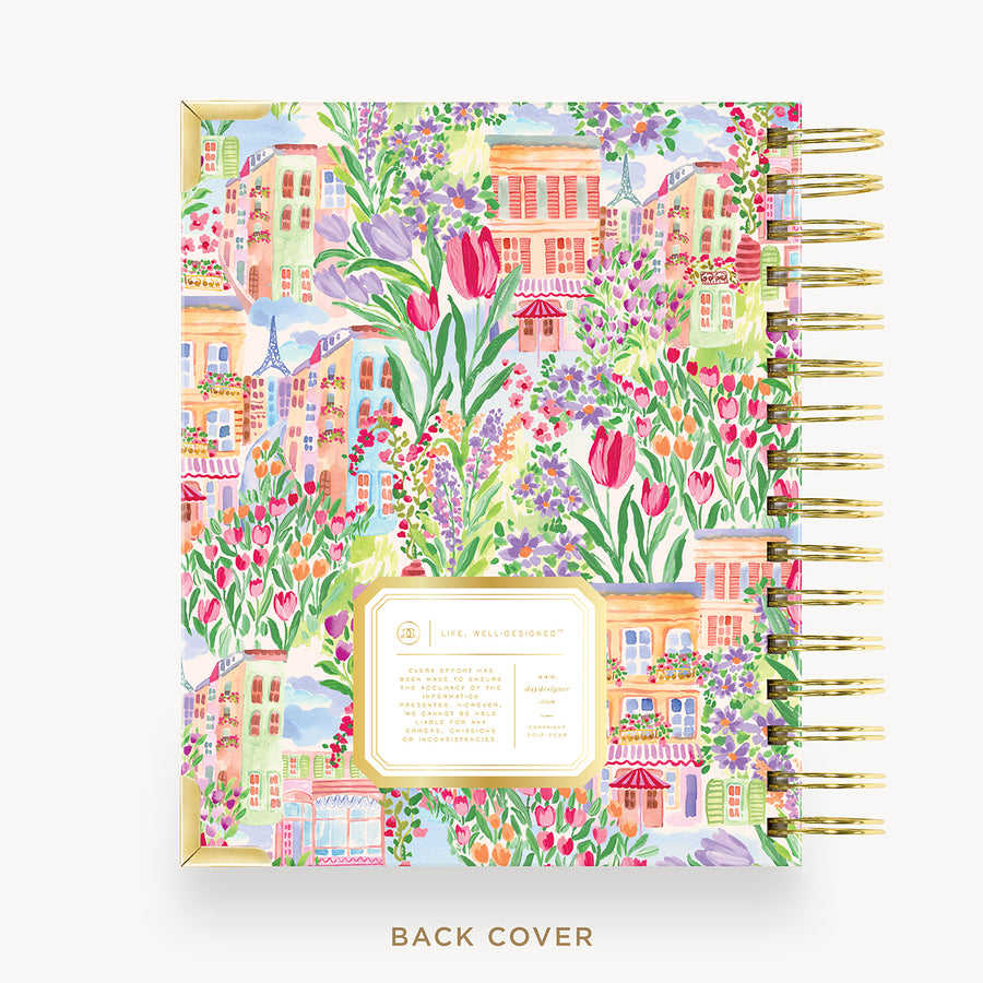 A spiral-bound notebook or planner shown from the back, featuring a colorful illustrated pattern of flowers, tulips, and small buildings. The cover has a cream label area with text, and the pages are bound with gold-tone spiral rings.