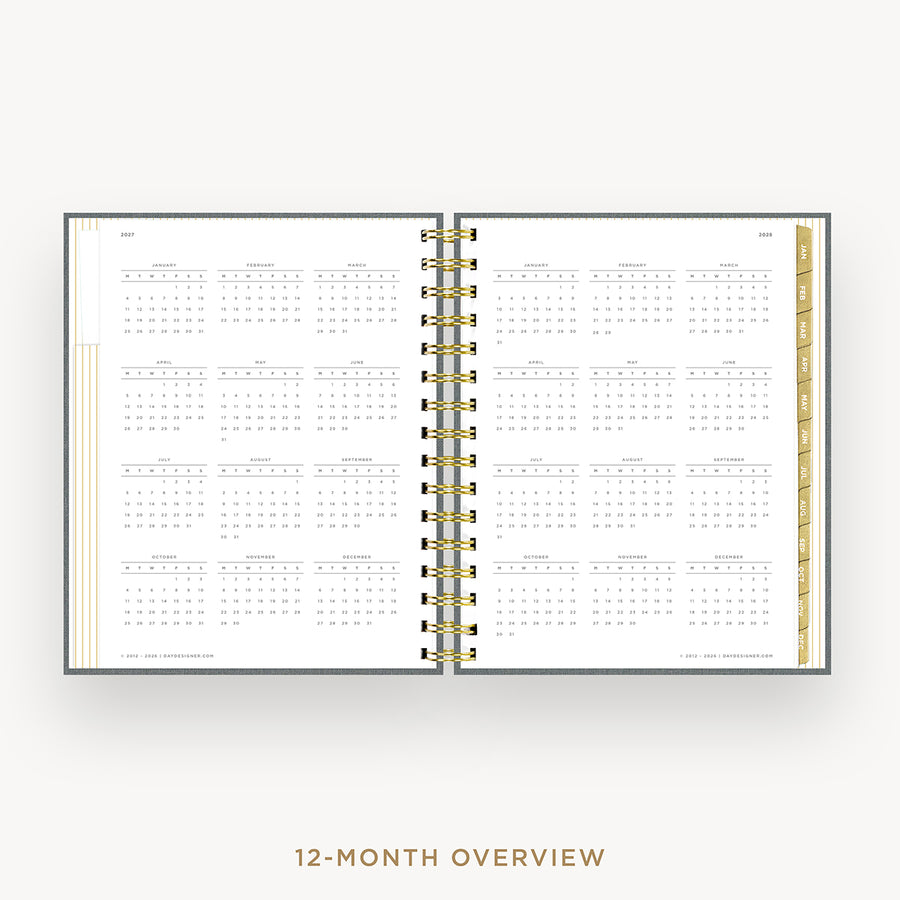 An open spiral-bound planner showing a 12-month calendar overview layout on white pages, with gold-toned ring binding and a gold vertical divider, centered on a light gray background with the text '12-MONTH OVERVIEW' below.