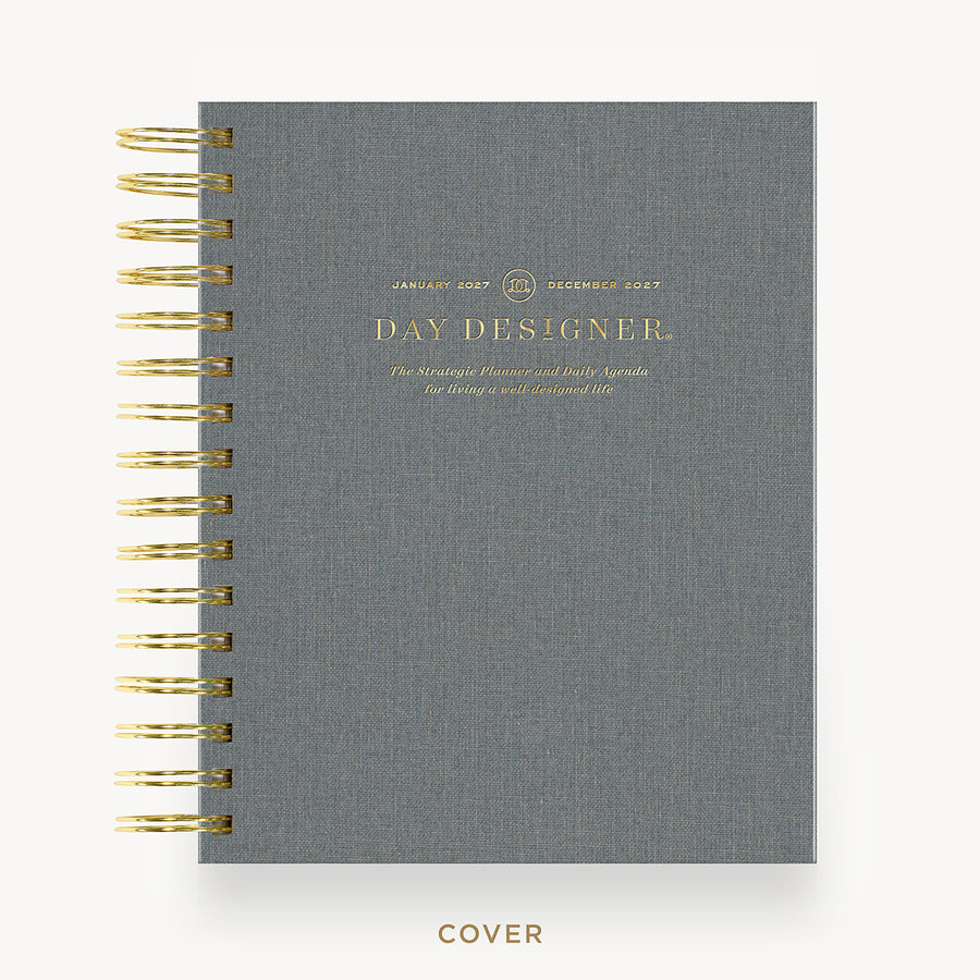 A gray linen-textured spiral notebook cover with gold wire binding and gold foil text reading 'DAY DESIGNER' on a white background.