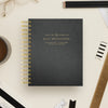 A black textured spiral-bound planner or day designer notebook with gold lettering, photographed from above on a light beige desk with pencils, glasses, a coffee cup, and other stationery props.