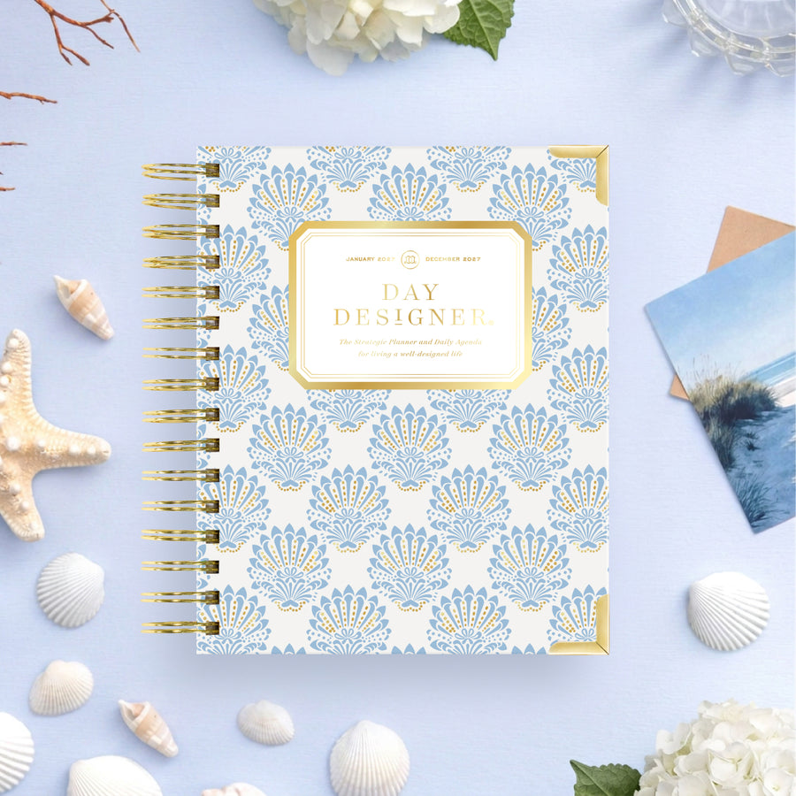 A spiral-bound planner or journal titled 'DAY DESIGNER' lies on a light blue surface with decorative seashells, flowers, and a small photo card around it. The cover has a repeating blue and white fan pattern with gold trim and a gold title panel.