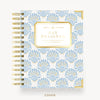 A Day Designer spiral-bound planner cover shown flat against a light gray background. The cover features a repeating light blue and gold damask pattern on a white base, with a gold-bordered central label reading 'DAY DESIGNER' and gold spiral binding along the left side.