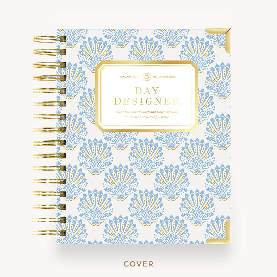 A Day Designer spiral-bound planner cover shown flat against a light gray background. The cover features a repeating light blue and gold damask pattern on a white base, with a gold-bordered central label reading 'DAY DESIGNER' and gold spiral binding along the left side.