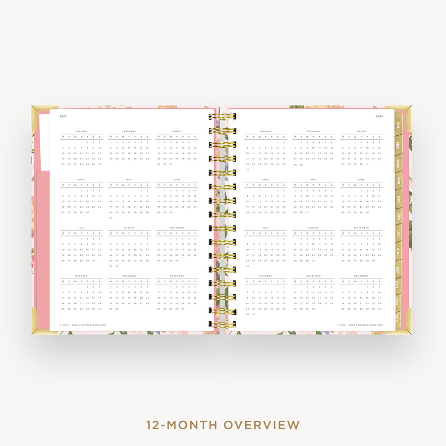 An open 12-month spiral-bound planner or calendar showing a monthly overview layout on white pages, with a pink cover edge and gold wire binding, centered on a light gray background with the text '12-MONTH OVERVIEW' below.