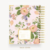 A floral spiral notebook shown from the back cover, featuring a light pink background with large pink and white flowers, green leaves, and purple blossoms. The cover has a gold border and a gold spiral binding on the right side.