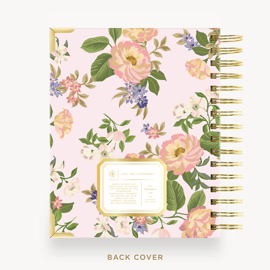 A floral spiral notebook shown from the back cover, featuring a light pink background with large pink and white flowers, green leaves, and purple blossoms. The cover has a gold border and a gold spiral binding on the right side.