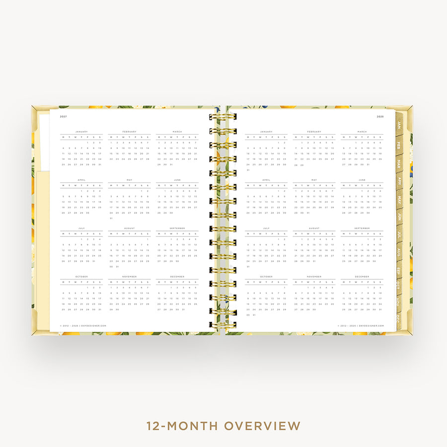 An open spiral-bound planner or calendar showing a 12-month overview layout on white pages, with a floral-patterned cover and gold spiral binding, centered on a light gray background with the text '12-MONTH OVERVIEW' below.