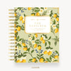 A Day Designer spiral-bound planner shown from the front on a light gray background. The cover features a botanical pattern with yellow lemons, white flowers, green leaves, and small blue berries on a pale sage green base.