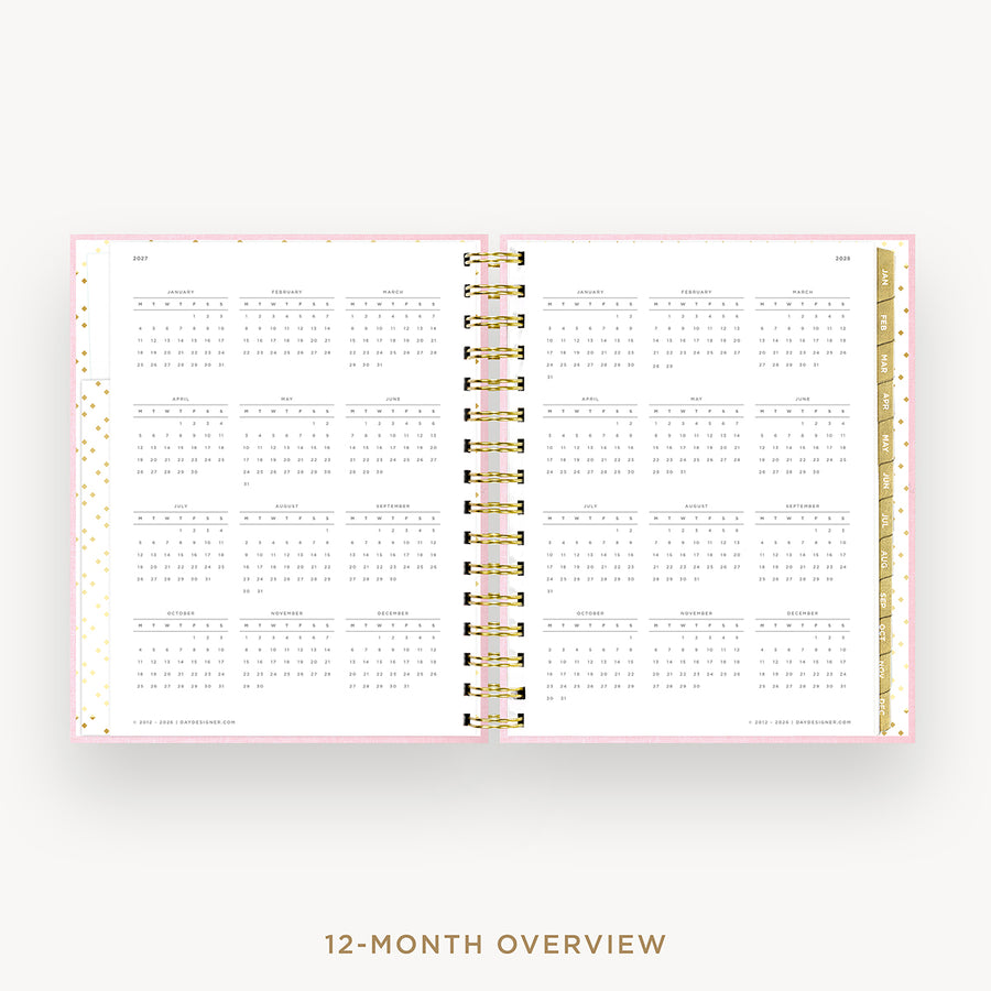 An open spiral-bound planner showing a 12-month calendar overview layout on white pages with a light pink border and gold accents, centered on a light gray background with the text '12-MONTH OVERVIEW' below.