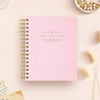 A pink spiral-bound planner or journal with gold lettering reading 'DAY DESIGNER' lies on a light beige surface with styled props including dried grass, beads, and paper notes.