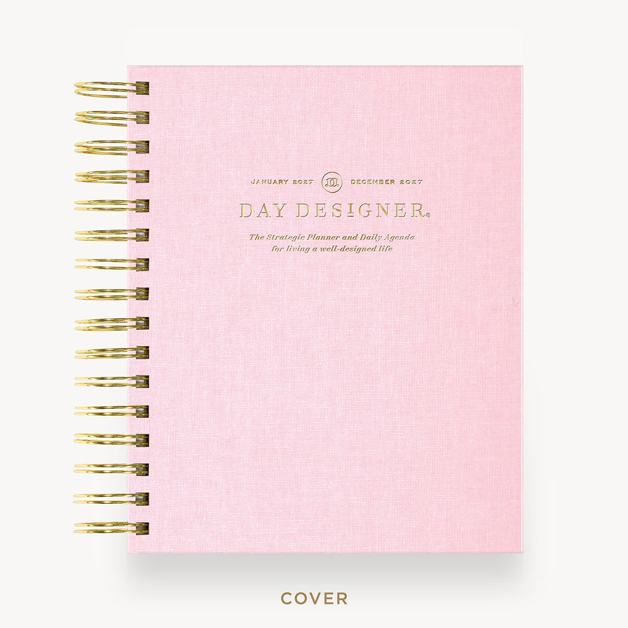A pink spiral-bound notebook cover with gold wire binding and gold foil text reading 'DAY DESIGNER.' on a clean white background.