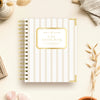 A spiral-bound planner titled 'DAY DESIGNER.' lies on a light cream surface with decorative props around it, including dried grass, crystals, a small vase, and folded fabric.