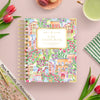 A spiral-bound planner titled 'DAY DESIGNER.' with a colorful floral and architectural illustration, styled in a flat lay on a pink surface with tulips, a cup of green tea, and a striped cloth.