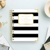 A Day Designer spiral notebook with bold black and white horizontal stripes and gold accents, styled on a light blue desk with office props.
