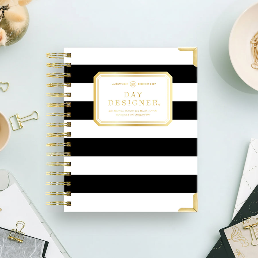 A Day Designer spiral notebook with bold black and white horizontal stripes and gold accents, styled on a light blue desk with office props.