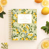 A Day Designer spiral-bound planner by Lemon & Clover sits on a light cream background, styled with fresh lemons, green leaves, and small yellow flowers. The planner cover features a light sage green background decorated with a lemon and white flower pattern, with a cream label in the center.