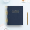 A navy blue spiral-bound planner or journal titled 'DAY DESIGNER.' lies on a light blue surface, styled with a gold pen, a glass bowl, and small paper props.