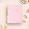 A pink textured spiral notebook with gold foil lettering that reads 'DAY DESIGNER.' It is photographed from above on a light beige surface with small decorative props around it, including dried grass, beads, and a handwritten note.