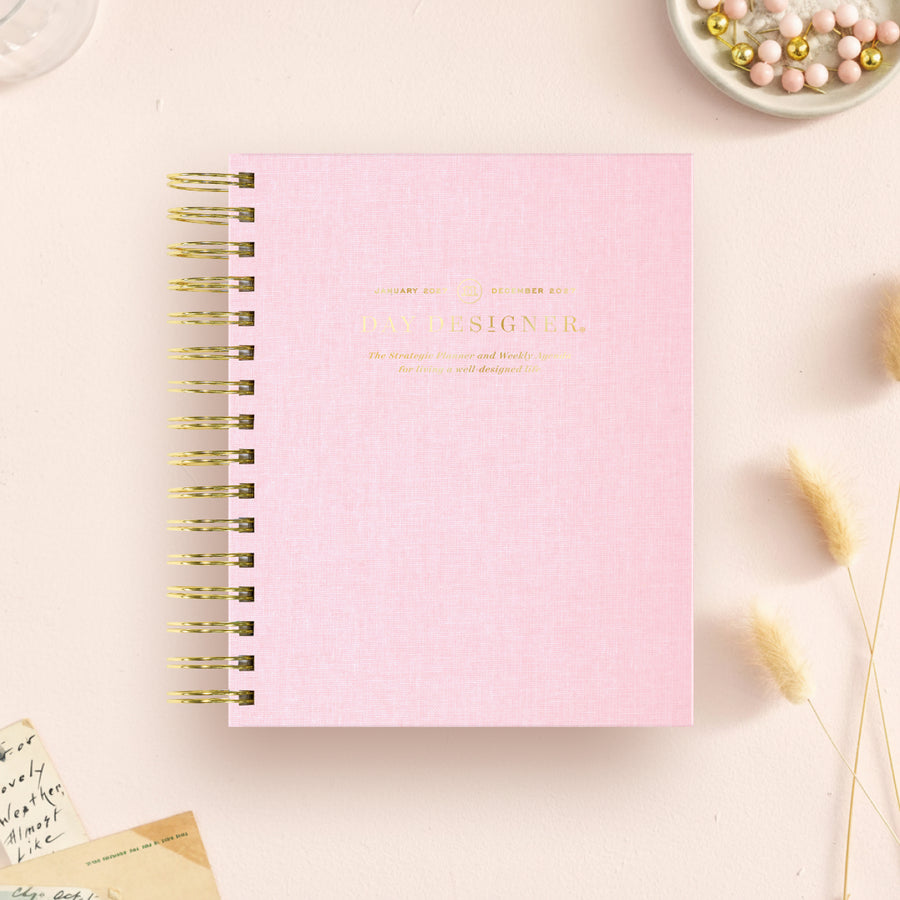 A pink textured spiral notebook with gold foil lettering that reads 'DAY DESIGNER.' It is photographed from above on a light beige surface with small decorative props around it, including dried grass, beads, and a handwritten note.