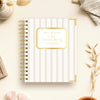 A spiral-bound planner titled 'DAY DESIGNER.' lies on a light cream surface with decorative props around it, including dried grass, crystals, a small vase, and folded fabric.
