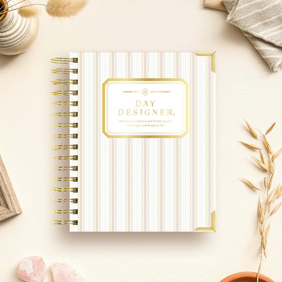 A spiral-bound planner titled 'DAY DESIGNER.' lies on a light cream surface with decorative props around it, including dried grass, crystals, a small vase, and folded fabric.