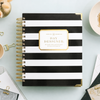 A spiral-bound planner or notebook with bold black and white horizontal stripes and a gold label reading 'DAY DESIGNER.' It is styled on a pale blue surface with gold paper clips, a coffee cup, and other desk accessories around it.