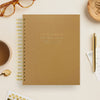 Brown notebook with gold spiral binding on a light surface with a cup of tea, glasses, and other items.