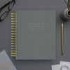 A gray spiral-bound day planner with gold text and gold wire binding, styled on a gray desk with a gold pen, glasses, and a small potted plant.