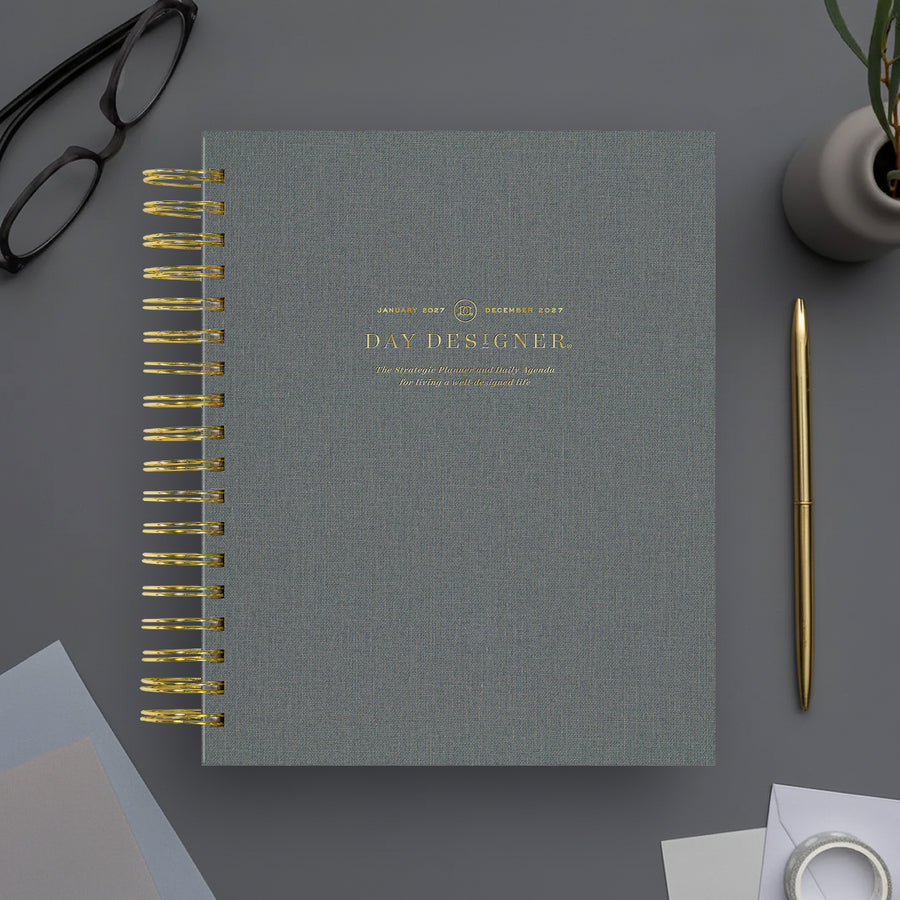 A gray spiral-bound day planner with gold text and gold wire binding, styled on a gray desk with a gold pen, glasses, and a small potted plant.