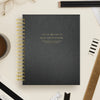 A black textured spiral-bound notebook with gold lettering reading 'DAY DESIGNER' and smaller text 'January 2027 | December 2027 is photographed from above on a light beige desk with pencils, glasses, a coffee cup, and other stationery around it.
