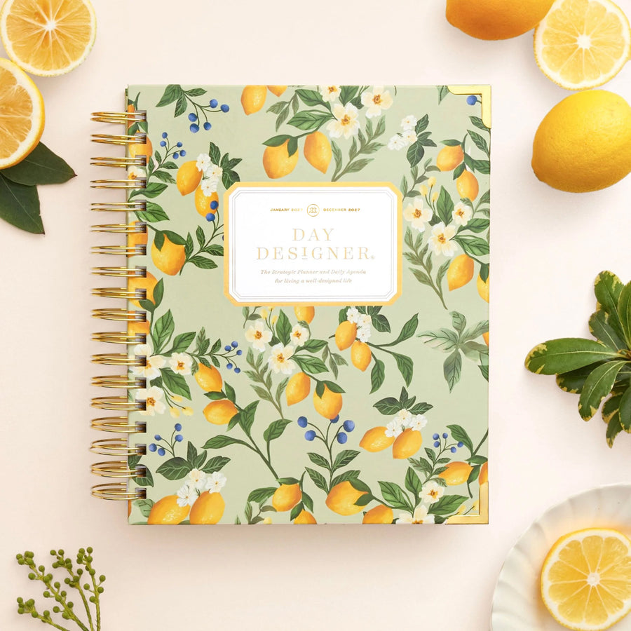 A spiral-bound daily planner with a light green cover decorated with a lemon and white flower pattern, styled in a flat lay with lemons and green leaves on a light beige background.