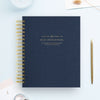A navy blue spiral-bound Day Designer notebook with gold lettering, styled in a flat-lay arrangement with a gold pen, paper clips, and a white background.