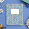 A spiral-bound planner or notebook titled 'DAY DESIGNER.' lies on a blue surface, surrounded by styled props including a glass dish, a small flower card, a brass object, and pages fanned out.